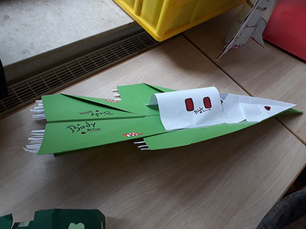 Ein handgefertigtes Modellflugzeug in leuchtendem Grün liegt auf einem Tisch, umgeben von anderen Bastelmaterialien.