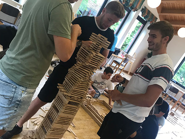 Eine Gruppe von Männern konstruiert einen beeindruckenden Holzstapel, umgeben von verstreuten Stäbchen auf dem Boden.