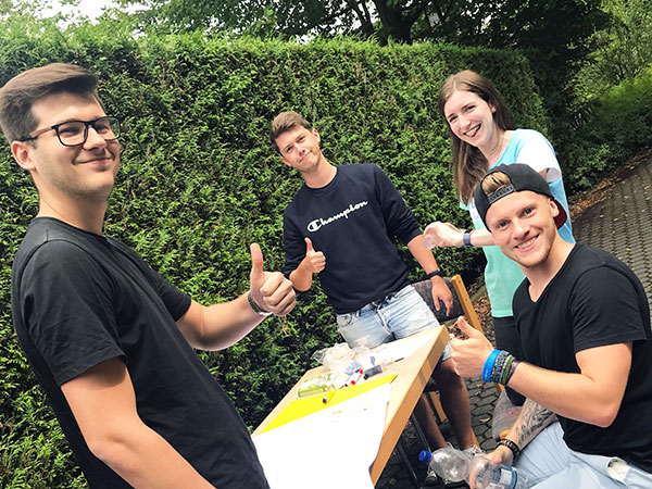 Vier junge Erwachsene stehen an einem Tisch im Freien und zeigen begeistert Daumen hoch, um ihre Freude auszudrücken.